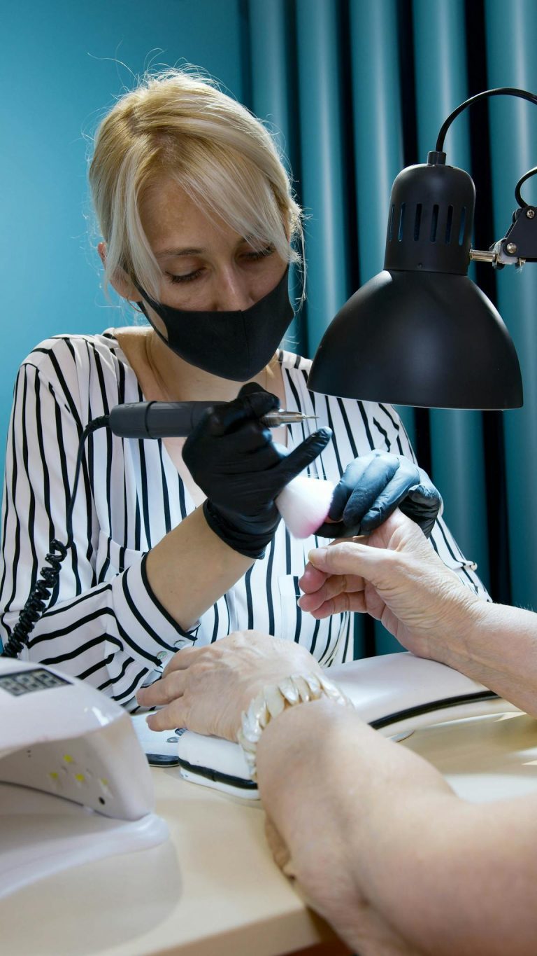 A manicurist wearing a face mask and gloves provides nail care service under a desk lamp.